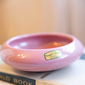 Op de afbeelding: Een roze keramische kom met een afgeronde vorm, rustend op een boek. De kom heeft een glanzende afwerking en een sticker met de tekst "Haeger Floral, Macomb, IL, Made in U.S.A.". Het boek heeft een donkergroene rug met gouden letters.
