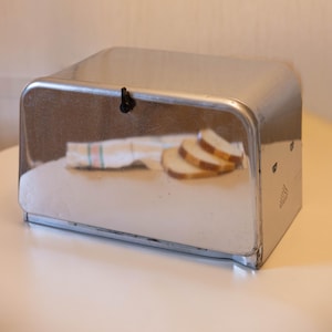 Op de afbeelding: Een glanzende, rechthoekige brooddoos van metaal met een gebogen bovenkant en een kleine zwarte sluiting. Binnenin een witte doek met groene en roze strepen en drie sneetjes brood.