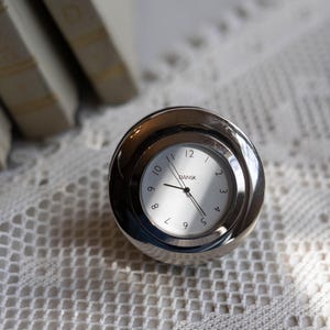 May include: A silver desk clock with a white face and black numerals. The clock, branded "DANSK", sits on a white, open-weave surface. The clock's hands indicate the time. Books are visible in the background.