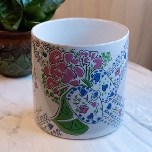 May include: White ceramic planter with a floral design in pink, blue, and green. The cylindrical pot features a detailed pattern of flowers and leaves, perfect for indoor plants. A small green plant is visible in the background.
