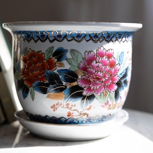 Op de afbeelding: Witte keramische bloempot met bijpassende schotel. De bloempot is versierd met oranje en roze bloemmotieven, groene bladeren en gouden accenten. De rand heeft een blauw-gouden rand.