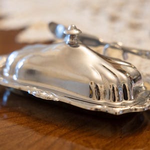 May include: A silver butter dish with a lid and a matching butter knife. The dish has a decorative, scalloped edge and a shiny, reflective surface. The butter dish is on a wooden surface.