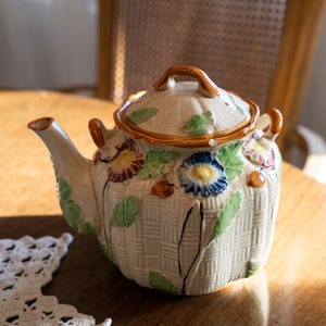 May include: Cream-colored ceramic teapot with a basket weave design. The teapot features colorful floral accents in blue, purple, and yellow, with green leaves. It has a brown handle and rim, and a matching lid. A charming addition to any tea set.