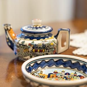 May include: A ceramic teapot and bowl set with a detailed, colorful cityscape design. The teapot features a blue and white lid and handle, with orange accents. The bowl has a similar design, with a blue wave pattern around the rim.