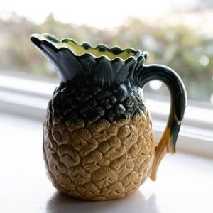 May include: A ceramic pitcher shaped like a pineapple. The pitcher has a textured, yellow body and a green top with a scalloped edge. The handle is green and yellow. The pitcher is sitting on a white surface.