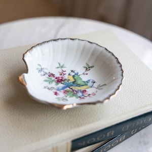 以下が含まれることがあります： 金色の縁取りが付いた、貝殻型の小さな白い皿。青と黄色の鳥がピンクの花と緑の葉の間に描かれています。皿は本の上に置かれています。