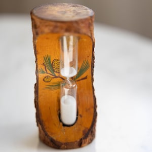 May include: A decorative hourglass with white sand inside a clear glass tube. The hourglass is set in a rustic wooden frame with a natural bark edge. The frame features a painted pinecone and branch design.