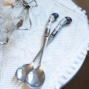 Peut inclure: Une paire de couverts à salade argentés avec des manches blancs ornés de motifs floraux complexes en bleu, or et multicolores. Les manches présentent des extrémités arrondies bleu foncé avec des détails dorés et des cuillerons polis.