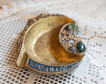 May include: A crescent-shaped gold-toned metal dish featuring the word JERUSALEM in blue lettering along the bottom edge. The dish includes intricate filigree detailing, small colorful accents, and a single oval-shaped green stone set on the right side.