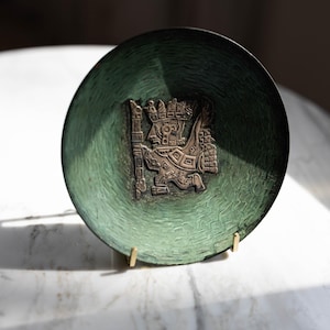 Könnte beinhalten: Eine runde, grüne Zierplatte mit einem detaillierten Relief einer Figur, die einen Stab hält. Die Platte hat eine strukturierte Oberfläche und wird auf einem goldfarbenen Ständer präsentiert. Die Platte ist wahrscheinlich aus Metall.