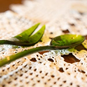 Puede incluir: Un juego de tenedor y cuchara de vidrio verde. Los cubiertos están sobre un mantel individual de encaje blanco, con una superficie de madera en el fondo. El tenedor tiene tres púas y la cuchara tiene un cuenco redondeado.