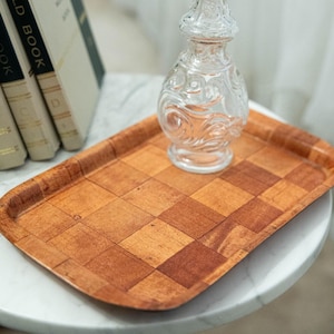 May include: A decorative wooden tray with a checkerboard pattern in shades of brown and tan. A clear glass bottle with an ornate design sits on the tray. The tray is on a white marble surface, with a stack of books in the background.