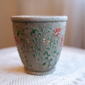 May include: A gray, ceramic plant pot with a floral design. The pot features a textured surface and is adorned with green vines, red and yellow flowers. The pot is sitting on a white, lace tablecloth.