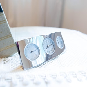 May include: A rectangular silver-toned desk accessory featuring three circular instruments: a hygrometer on the left, an analogue clock with Roman numerals in the centre, and a smaller circular gauge on the right. The surface is polished and reflective.