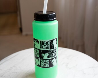 Puede incluir: Una botella de agua verde con tapa negra y pajita blanca. La botella tiene una pegatina con el texto "NEW KIDS ON THE BLOCK" e imágenes de los miembros de la banda. La botella está sobre una superficie de mármol blanco.