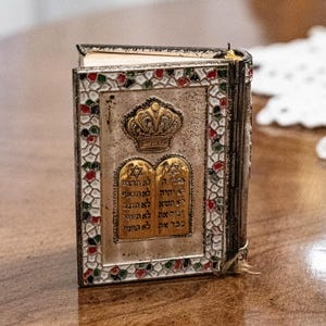 May include: A small, silver-toned book with a decorative cover. The cover features a gold crown and two tablets with Hebrew text. The border is adorned with red, green, and white accents. The book is standing upright on a wooden surface.