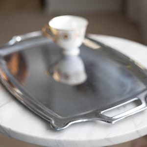 Puede incluir: Una bandeja plateada con asas sobre una mesa de mármol blanco. Una taza de té blanca con asa dorada y diseño floral descansa sobre la bandeja. La bandeja refleja la imagen de la taza.