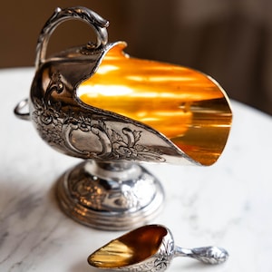 May include: Ornate silver sugar bowl with a matching scoop. The bowl has a gold-colored interior and is decorated with floral patterns. The scoop is small and has a similar design. The set is placed on a white marble surface.