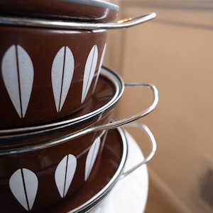 May include: Stacked brown enamelware pots with white leaf-shaped designs. Each pot has a silver handle and rim. The lids are also brown with silver rims. The pots are on a white surface.