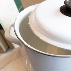 May include: A white enamel pot with a lid, featuring a black knob. The pot has a silver rim and handles. The lid is slightly ajar, revealing the interior. Books are visible in the background.