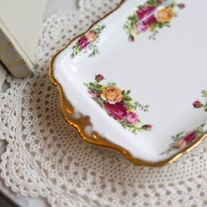 May include: A white rectangular serving tray with a gold rim and floral rose pattern. The tray is decorated with yellow, pink, and red roses. The tray is on a white lace doily.
