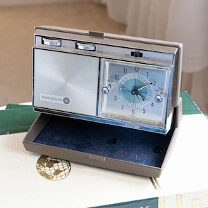 May include: A vintage Westinghouse clock radio with a brown exterior and silver accents. The clock face has a blue and silver design with black hands. The radio has a speaker, volume and tune dials, and an alarm function. The clock is open, revealing a dark blue interior.
