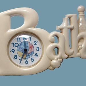 May include: Cream-colored "Bath" clock with a round clock face. The clock face has black numbers and hands. The word "Bath" is stylized with a bottle and bubbles. The clock is a decorative item.