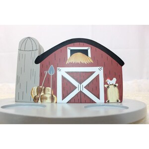 May include: A painted wooden barn-shaped stand featuring a red barn with white trim, a gray silo, and a black roof. The design includes a hayloft, a shovel, hay bales, and a small white chicken. The base is a light gray circular platform.