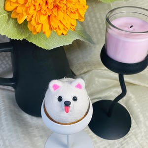 May include: A white felted dog ornament with pink ears and a red tongue, set in a white holder with a gold rim. The ornament is on a white stand, next to a pink candle in a glass jar and a black candle holder. Orange flowers are in a black vase.