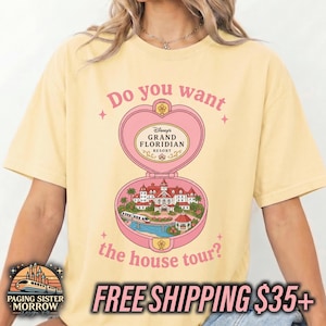 Puede incluir: Una camiseta de manga corta de color amarillo p&aacute;lido con un gr&aacute;fico de polvera rosa en forma de coraz&oacute;n. El dise&ntilde;o incluye el texto "Do you want the house tour?" y una ilustraci&oacute;n del Disney Grand Floridian Resort con un monorra&iacute;l y palmeras.