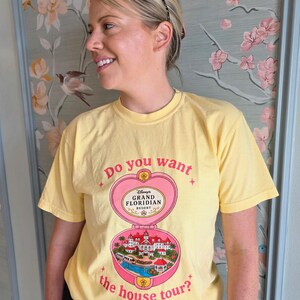 Puede incluir: Una camiseta de manga corta de color amarillo p&aacute;lido con un gr&aacute;fico de polvera rosa en forma de coraz&oacute;n. El texto dice Do you want the house tour? e incluye una ilustraci&oacute;n detallada del Disney's Grand Floridian Resort en el dise&ntilde;o.