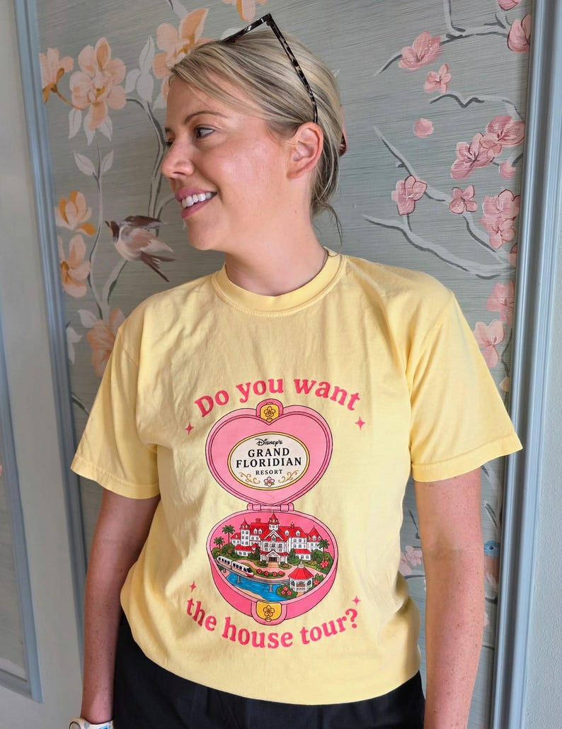 Puede incluir: Una camiseta de manga corta de color amarillo p&aacute;lido con un gr&aacute;fico de polvera rosa en forma de coraz&oacute;n. El texto dice Do you want the house tour? e incluye una ilustraci&oacute;n detallada del Disney's Grand Floridian Resort en el dise&ntilde;o.