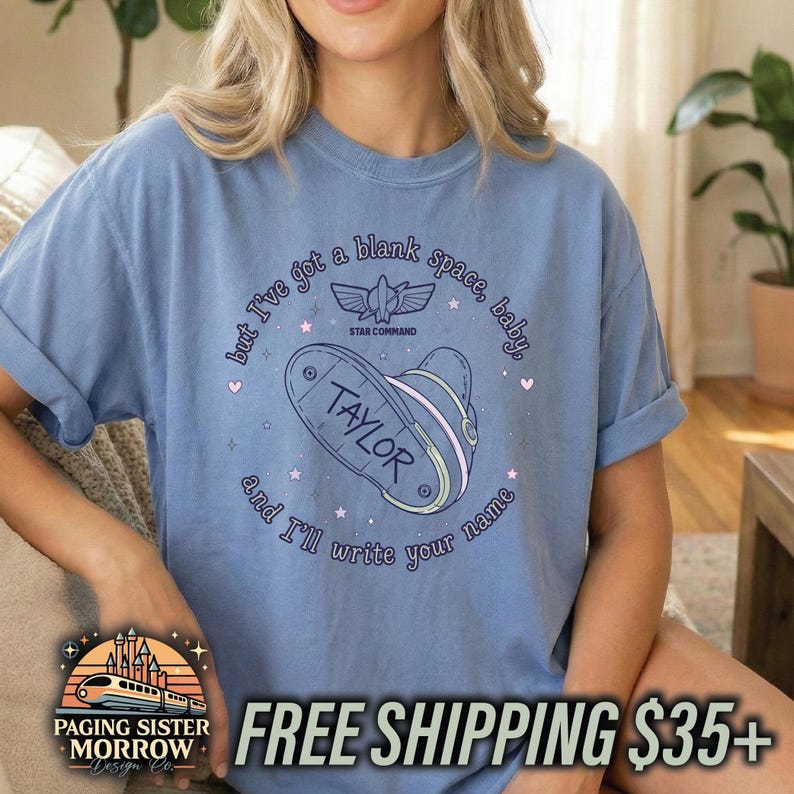 May include: A light blue short-sleeve t-shirt featuring a graphic print with the text "but I've got a blank space, baby, and I'll write your name" surrounding a stylised space helmet with the name "TAYLOR" and a "STAR COMMAND" logo.
