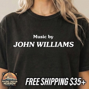 May include: Black t-shirt with the text "Music by JOHN WILLIAMS" in white. The shirt features a small logo at the bottom left and the text "FREE SHIPPING $35+" at the bottom right.