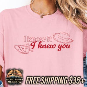 May include: A light pink t-shirt featuring the red text I knew it I knew you, accompanied by a line art illustration of a cowboy hat and a spur with the letters TS. The design is centered on the chest of the garment, which has a crew neckline.
