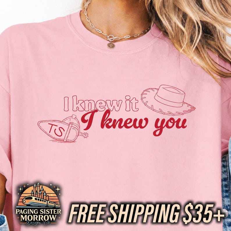 May include: A light pink t-shirt featuring the red text I knew it I knew you, accompanied by a line art illustration of a cowboy hat and a spur with the letters TS. The design is centered on the chest of the garment, which has a crew neckline.