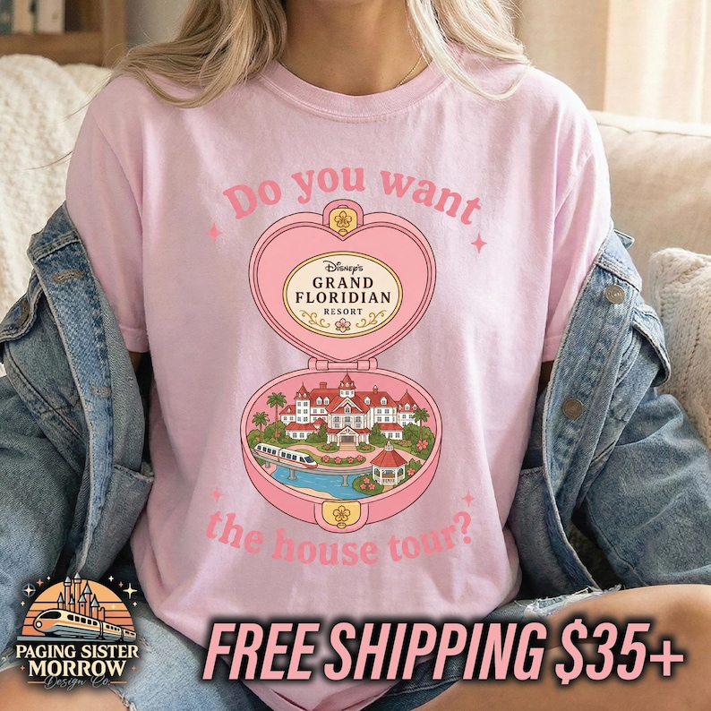 Puede incluir: Una camiseta rosa claro con un gr&aacute;fico de un estuche en forma de coraz&oacute;n que se abre para mostrar el Disney Grand Floridian Resort. El texto dice Do you want the house tour? El dise&ntilde;o incluye un monorra&iacute;l, palmeras y edificios.
