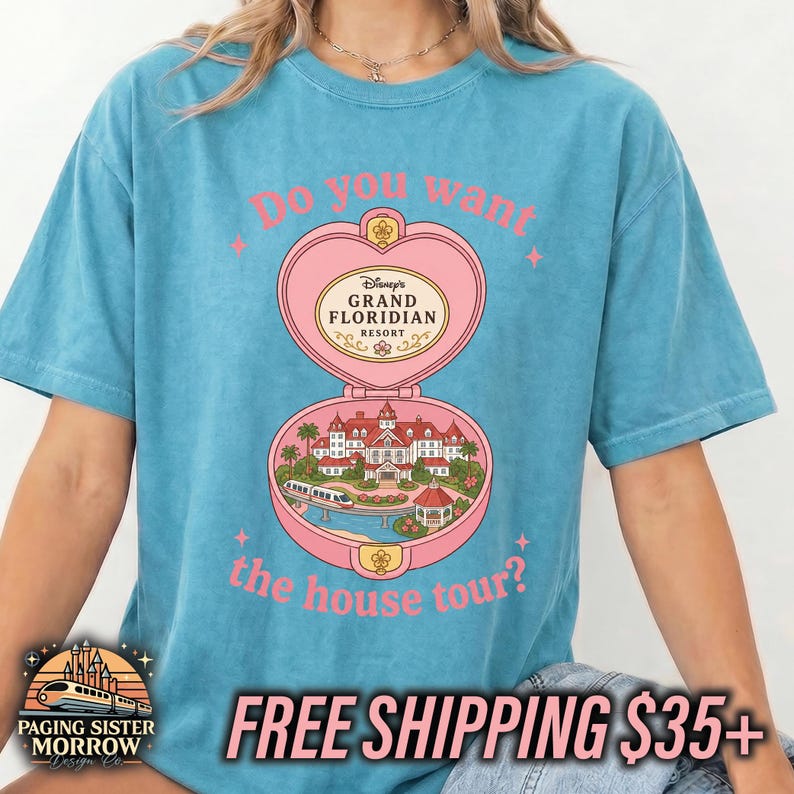 Puede incluir: Una camiseta azul de manga corta con un gr&aacute;fico de un estuche rosa en forma de coraz&oacute;n. El dise&ntilde;o muestra el Grand Floridian Resort con un monorra&iacute;l y el texto "Do you want the house tour?" en letras rosas encima y debajo de la ilustraci&oacute;n.