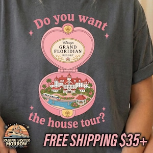 Puede incluir: Una camiseta gris carb&oacute;n con un gr&aacute;fico de un estuche compacto rosa en forma de coraz&oacute;n. Dentro hay una ilustraci&oacute;n del Disney Grand Floridian Resort con un monorra&iacute;l. El texto dice: Do you want the house tour? y Disney's Grand Floridian Resort.