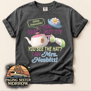 May include: Dark gray t-shirt with a whimsical design featuring a teapot, a blue hat with a flower, and the text "DON'T YOU GET IT? YOU SEE THE HAT? I AM Mrs. Nesbitt!". The shirt also includes the text "Ohhh Hannahh!" and the "PAGING SISTER MORROW Design Co." logo.