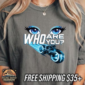 May include: Gray t-shirt featuring the text "WHO ARE YOU?" above a neon blue motorcycle graphic. The design includes blue eyes with long eyelashes. The shirt is a comfortable, casual style.