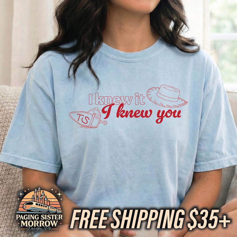 May include: A light blue short-sleeve t-shirt featuring red line art of a cowboy hat and a guitar pick with the letters TS. The text I knew it I knew you is printed in red across the center of the chest in a stylized script font.