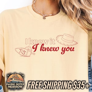 May include: A pale yellow t-shirt featuring the red text I knew it I knew you. The design includes a red outline of a cowboy hat and a spur with the letters TS. The shirt has a crew neckline and a relaxed fit, showcasing a graphic print.