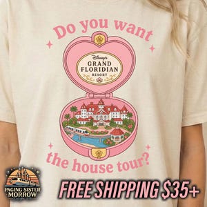 Puede incluir: Una camiseta beige con un gr&aacute;fico de un estuche rosa en forma de coraz&oacute;n. La parte superior muestra el texto "Do you want" y "Disney's GRAND FLORIDIAN RESORT". La parte inferior ilustra el complejo hotelero, un monorra&iacute;l y "the house tour?"