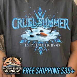 May include: Dark gray t-shirt featuring the text "CRUEL SUMMER" in light blue, with a whimsical illustration of a melting snowman. The design includes a carrot nose, stick arms, and black stone eyes. The phrase "THE SHAPE OF YOUR BODY, IT'S NEW" is below the illustration.