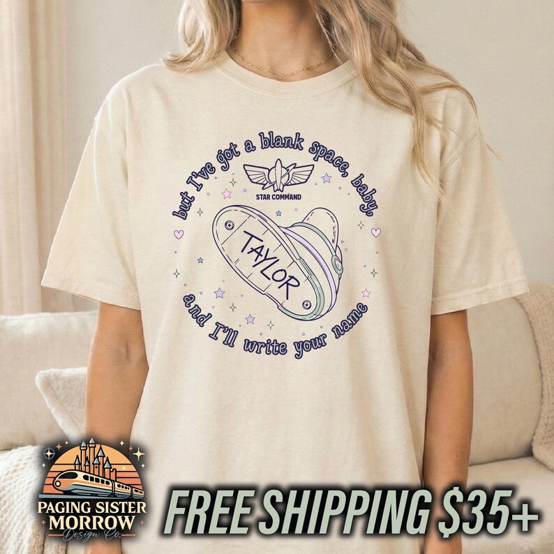 May include: A cream-coloured short-sleeve t-shirt featuring a graphic of a space ranger badge with the name Taylor. The text reads, but I've got a blank space, baby, and I'll write your name, surrounding the badge with small stars and hearts.