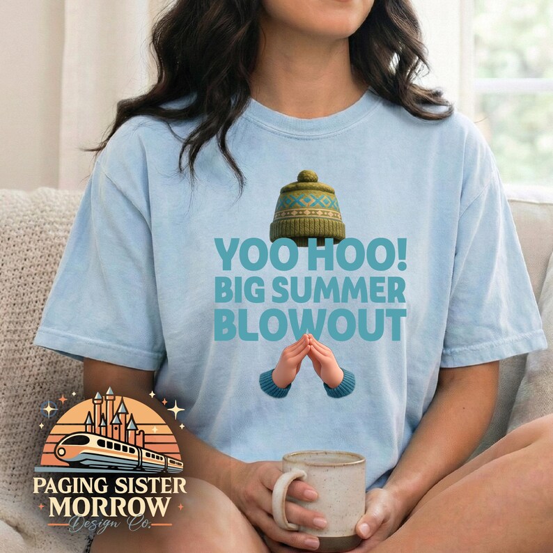 May include: Light blue t-shirt with the text "YOO HOO! BIG SUMMER BLOWOUT" and a graphic of a knit hat and hands. The shirt also features a logo with a train and castle design. A person is holding a beige mug.