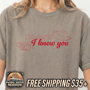 May include: A taupe short-sleeved t-shirt featuring red line art graphics of a cowboy hat and a spur with the letters TS. The text I knew it I knew you is printed in red across the center of the chest in a mix of block and script fonts.