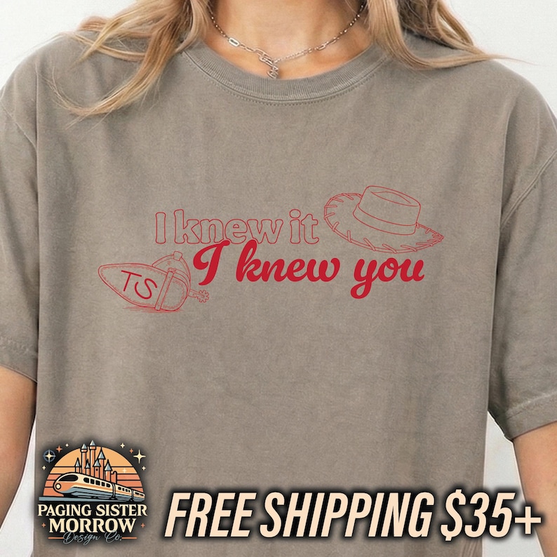 May include: A taupe short-sleeved t-shirt featuring red line art graphics of a cowboy hat and a spur with the letters TS. The text I knew it I knew you is printed in red across the center of the chest in a mix of block and script fonts.