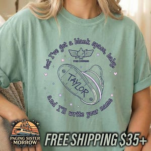 May include: A sage green short-sleeve t-shirt featuring a graphic print with the text "but I've got a blank space, baby, and I'll write your name" surrounding a space ranger helmet with the name "TAYLOR" and a "STAR COMMAND" logo.